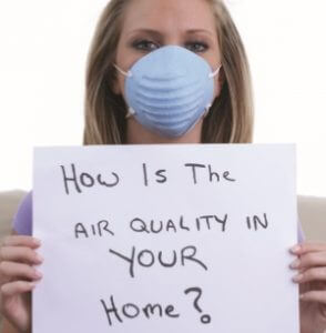 photo of a woman with blond hair and a mask on holding a sign that says: How is the air quality in YOUR home?